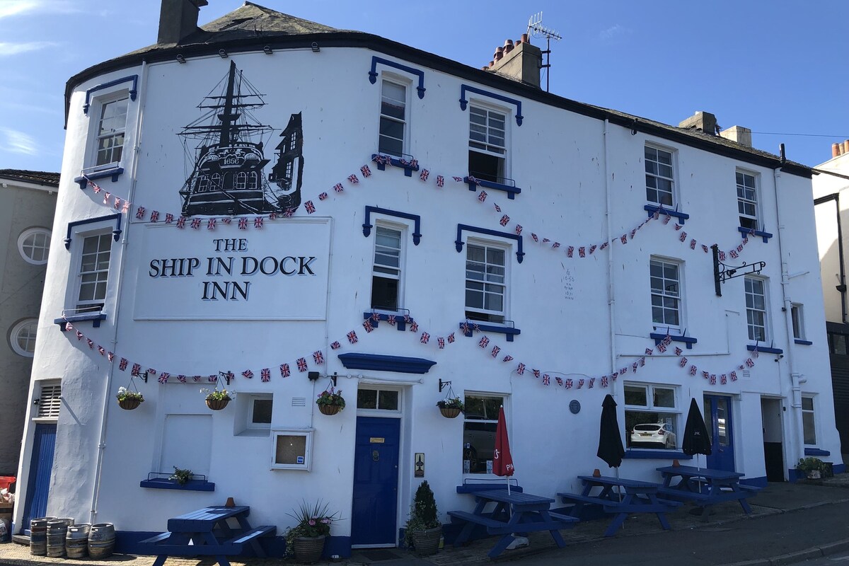 En hvid bygning med et skilt, der siger "The Ship in Dock Inn". Den har flere vinduer, en blå dør og udendørs siddepladser med parasoller.