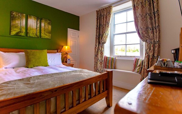 A hotel room with a bed, a bedside table with a lamp, a window with curtains, and a view of greenery outside.