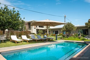 A swimming pool with lounge chairs and an umbrella, surrounded by greenery and buildings. - Schulphoek House (Hermanus)
