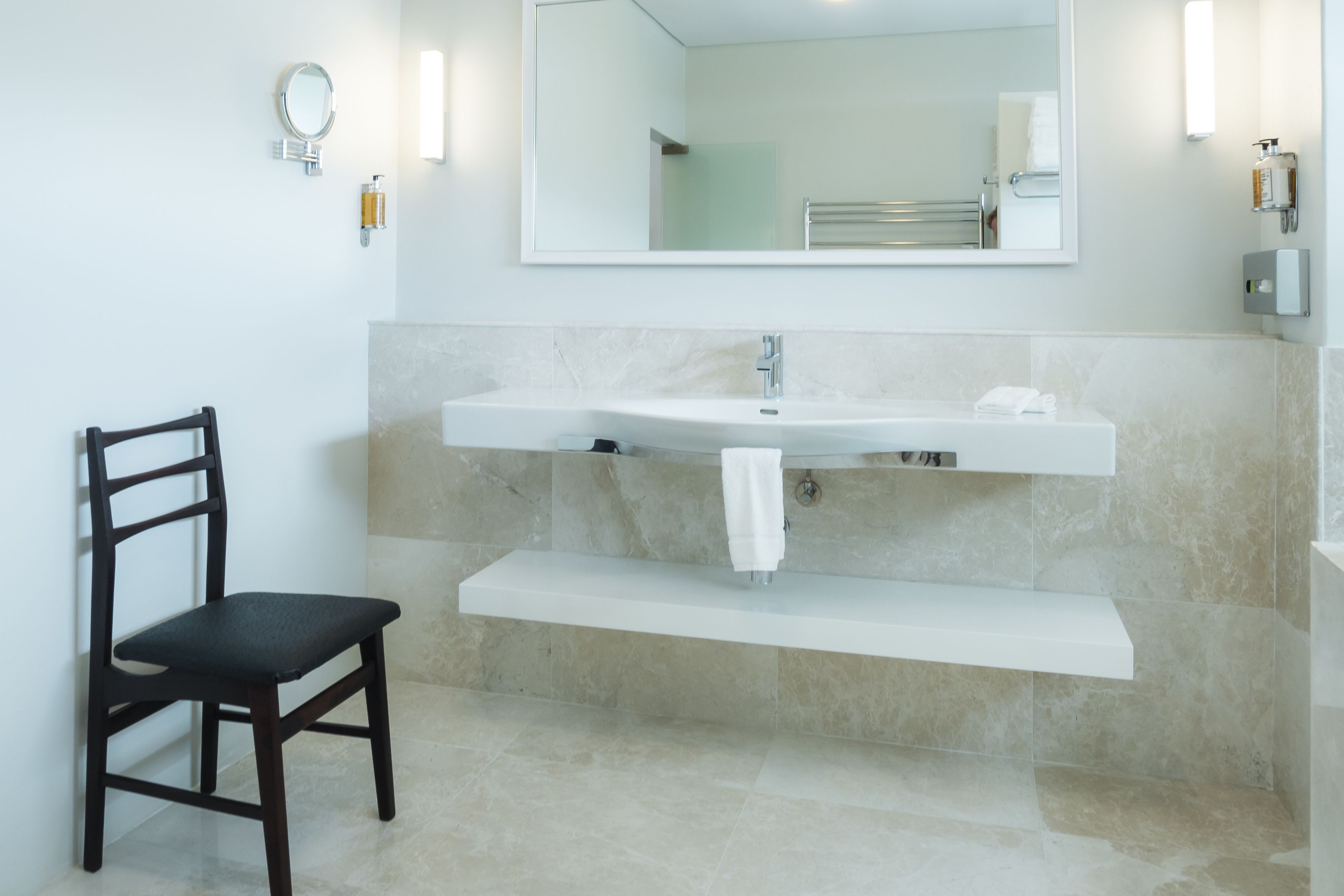 Un baño con lavamanos blanco, un espejo, toallero y una silla.
