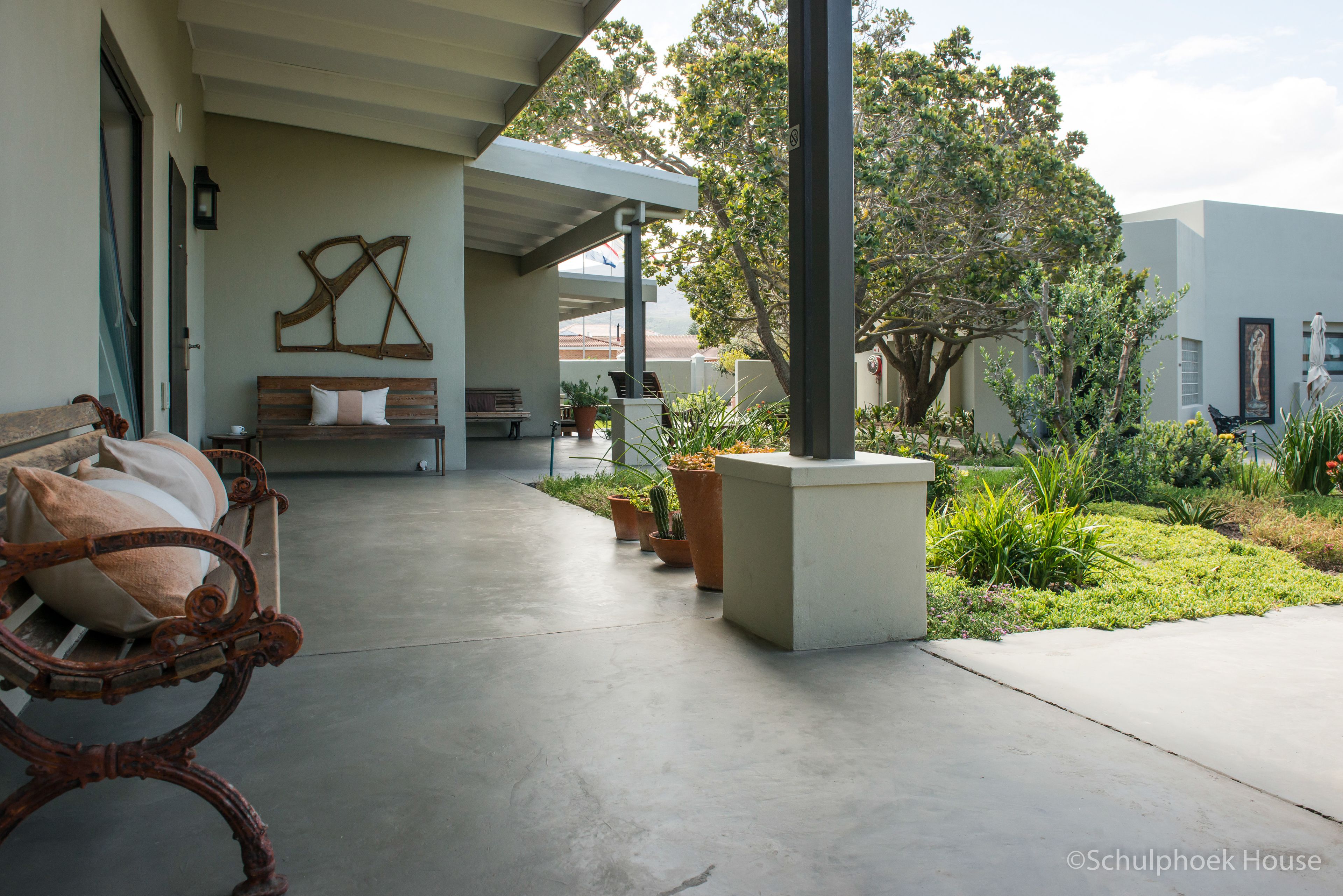 A covered outdoor seating area with a bench and a table, surrounded by potted plants and a tree.