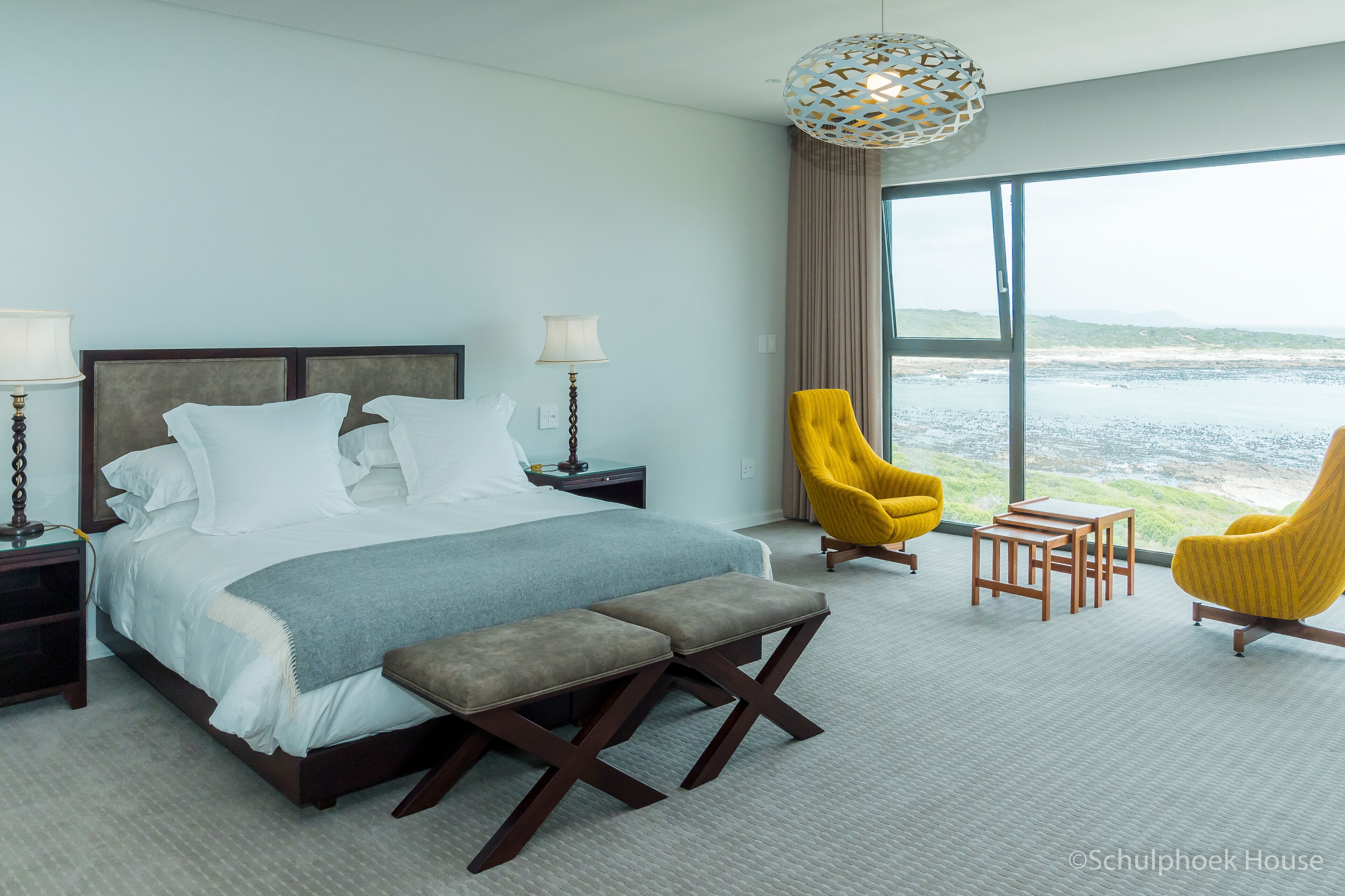 A modern hotel room with a large bed, two bedside tables, a bench, two yellow armchairs, and a view of the sea.
