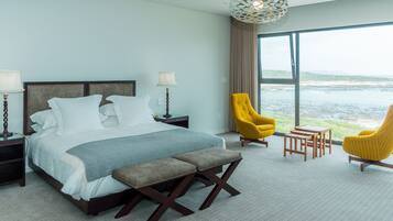 A modern hotel room with a large bed, two bedside tables, a bench, two yellow armchairs, and a view of the sea.