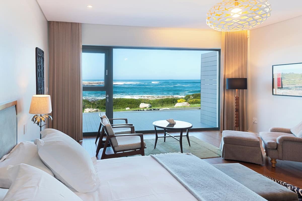 A hotel room with a large balcony, a bed, two armchairs, a small table, and a lamp.