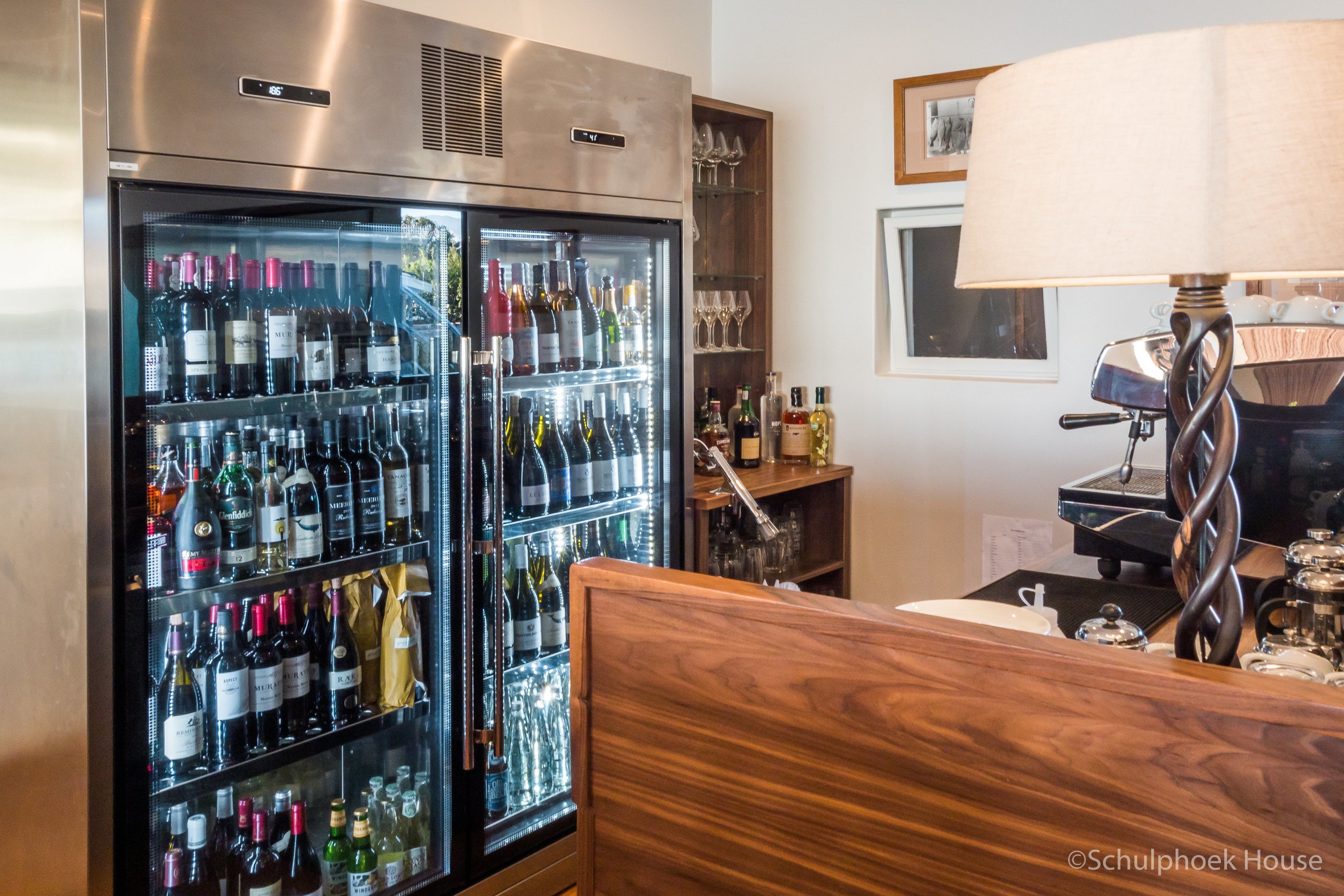 Habitación de hotel con minibar, cabecera de madera, cafetera y un refrigerador de vinos con varias botellas.