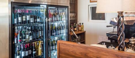 Habitación de hotel con minibar, cabecera de madera, cafetera y un refrigerador de vinos con varias botellas.