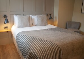 Uma cama bem arrumada com manta listrada, duas luminárias de cabeceira e uma cadeira em quarto de hotel.