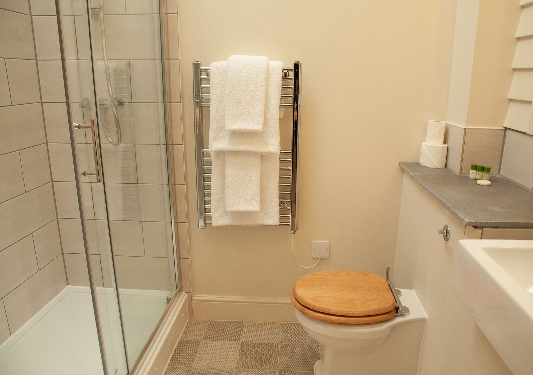 A bathroom with a glass shower enclosure, a white toilet with a wooden seat, and a towel rack with two white towels.