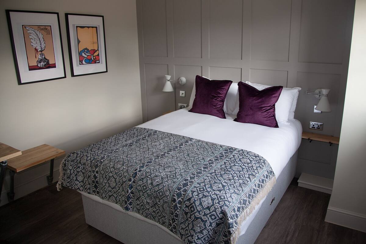 A neatly made bed with a patterned bedspread and two pillows, a bedside table with a lamp, and two framed artworks on the wall.