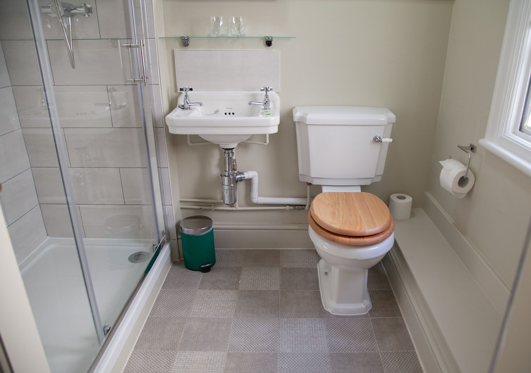 Une salle de bain avec une douche à parois de verre, un lavabo blanc, des toilettes avec un abattant en bois et une poubelle verte.