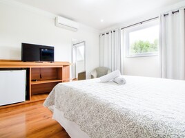 Quarto de hotel com cama, televisão em prateleira de madeira, mini-frigorífico e janela com cortinas.