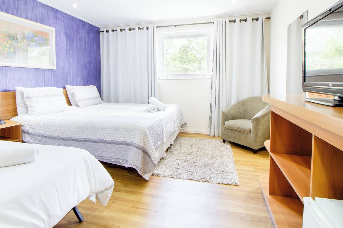 Une chambre d’hôtel avec un lit, une chaise, un bureau avec une télévision et une fenêtre avec des rideaux.