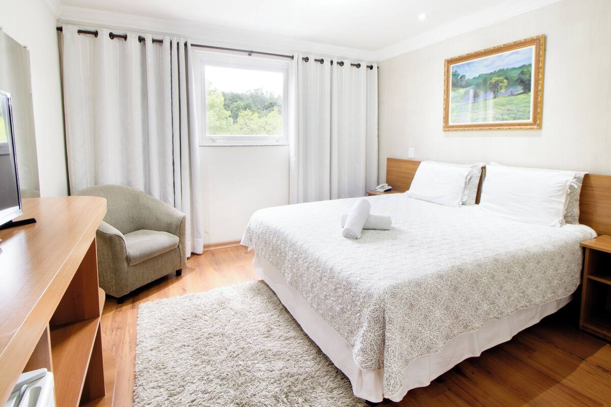 Une chambre d’hôtel avec un lit, un bureau, une chaise, une fenêtre avec des rideaux et un tableau au mur.