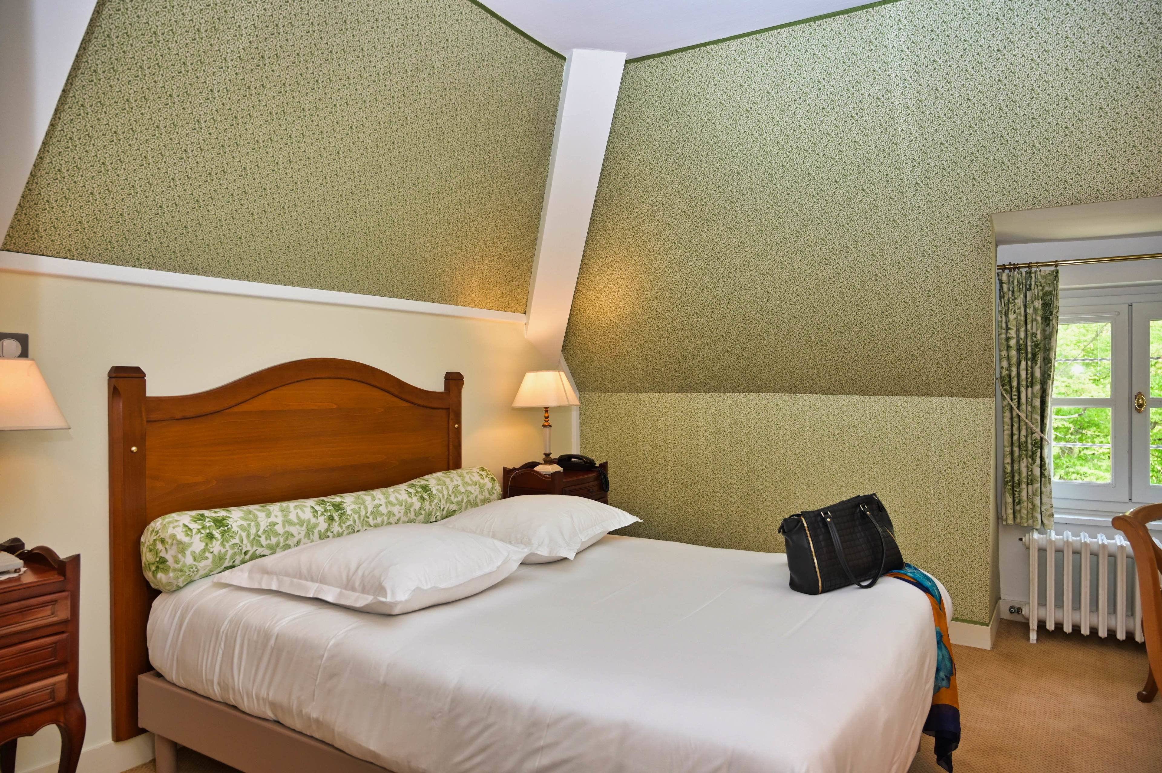 A neatly made bed with white linens and a green floral pillow, a wooden nightstand with a lamp, and a window with curtains.