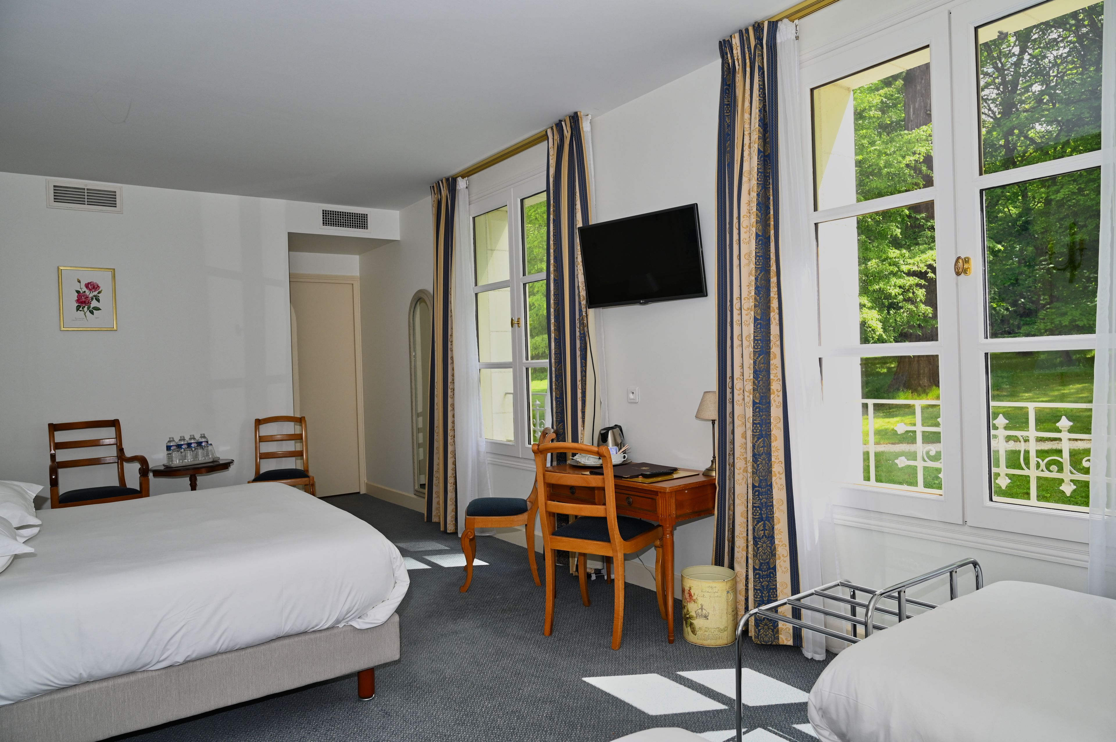 A hotel room with two beds, a desk with a chair, a television, and large windows with a view of greenery.