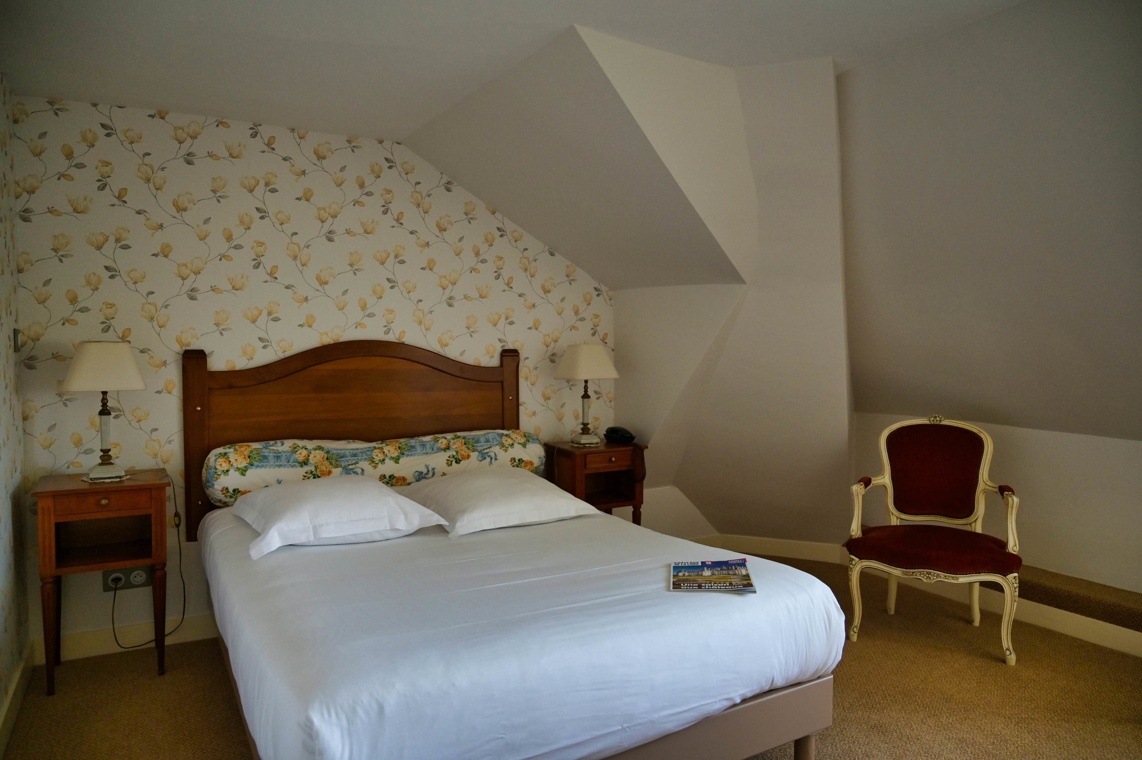 A neatly made bed with white linens, two bedside tables with lamps, a floral-patterned headboard, and a red upholstered chair.