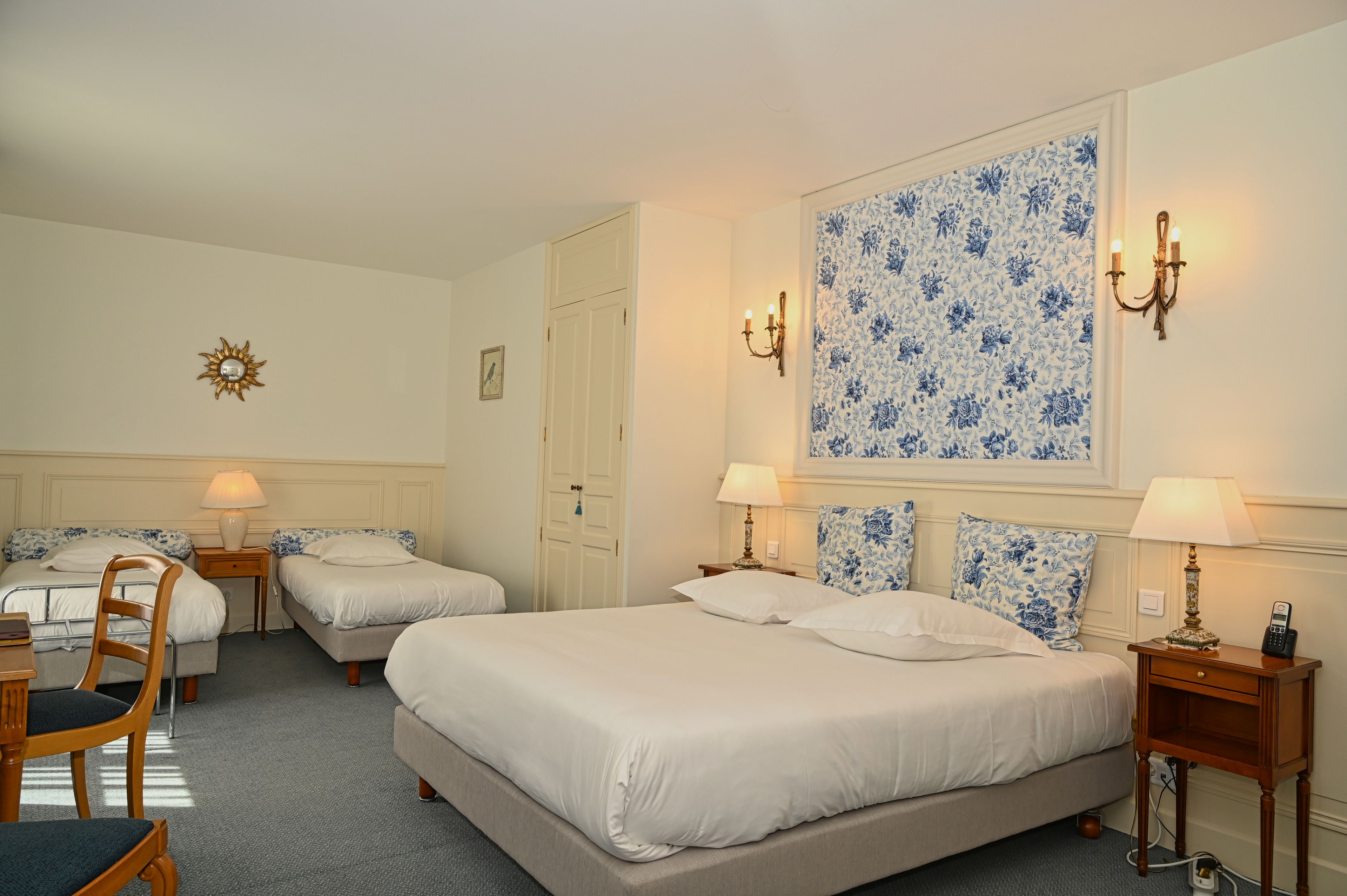 Une chambre d’hôtel avec deux lits simples, un bureau et une chaise. Il y a deux tables de chevet avec des lampes, ainsi qu’une lampe fixée au mur.