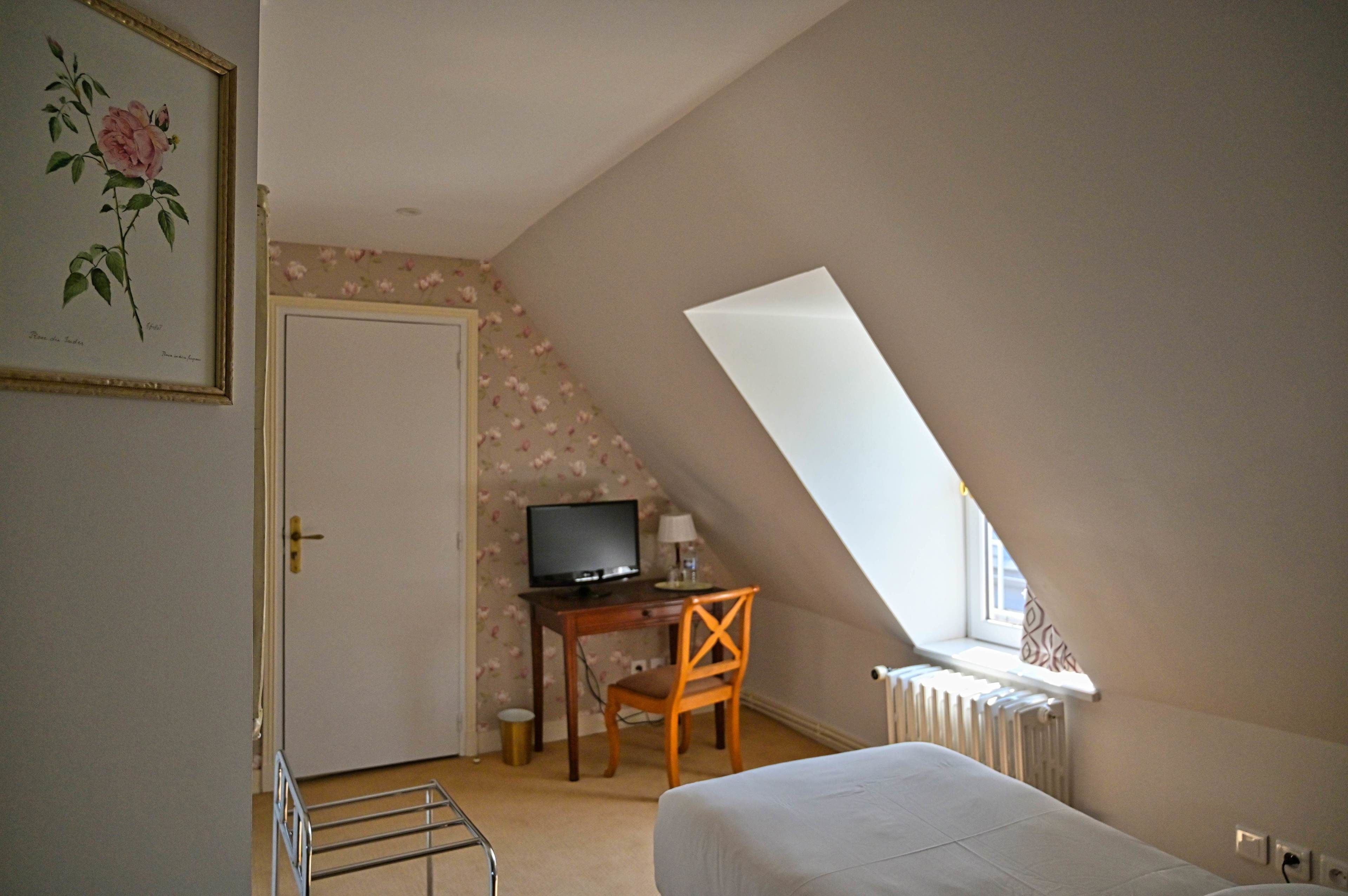 Un dormitorio con techo inclinado, una cama, un escritorio con silla, un televisor y un radiador.