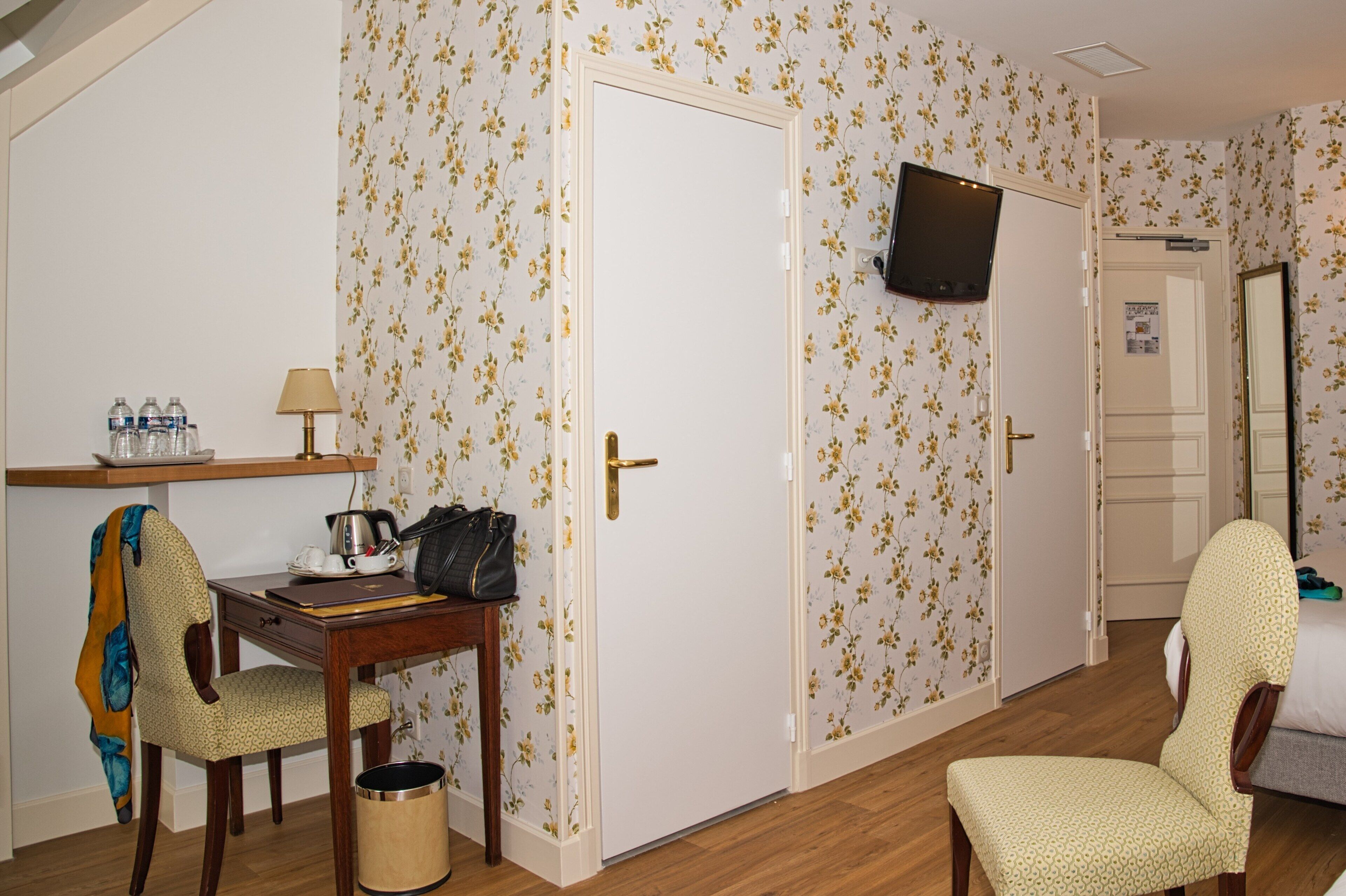 A hotel room with floral wallpaper, a wooden desk with a lamp, a chair, a bed, and a television mounted on the wall.