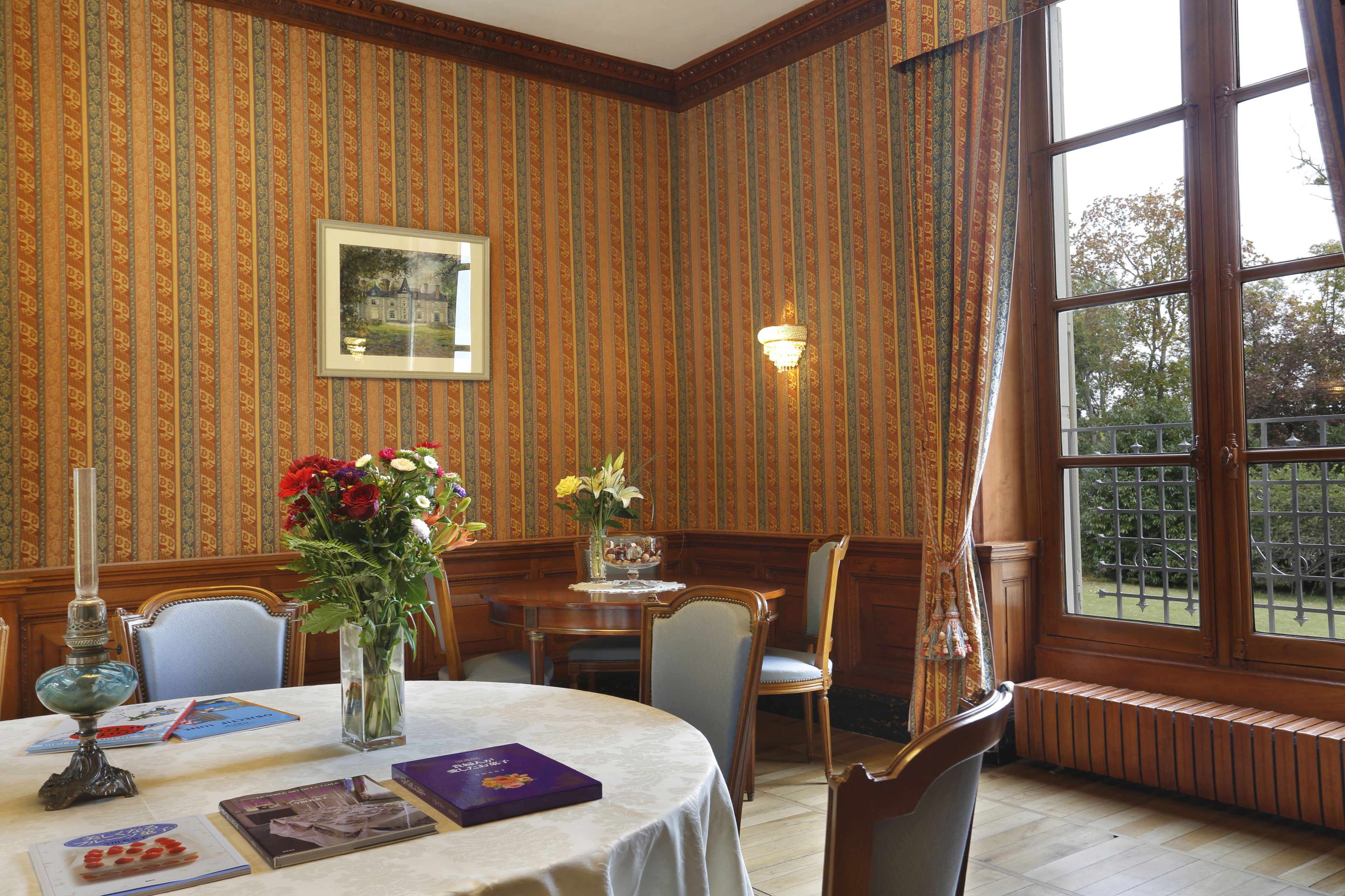 A dining room with a round table, chairs, a vase with flowers, a framed picture on the wall, and a window with a view of trees.