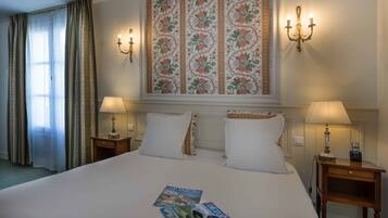 A hotel room with a large bed, two bedside tables with lamps, a wall-mounted light fixture, and a window with curtains.
