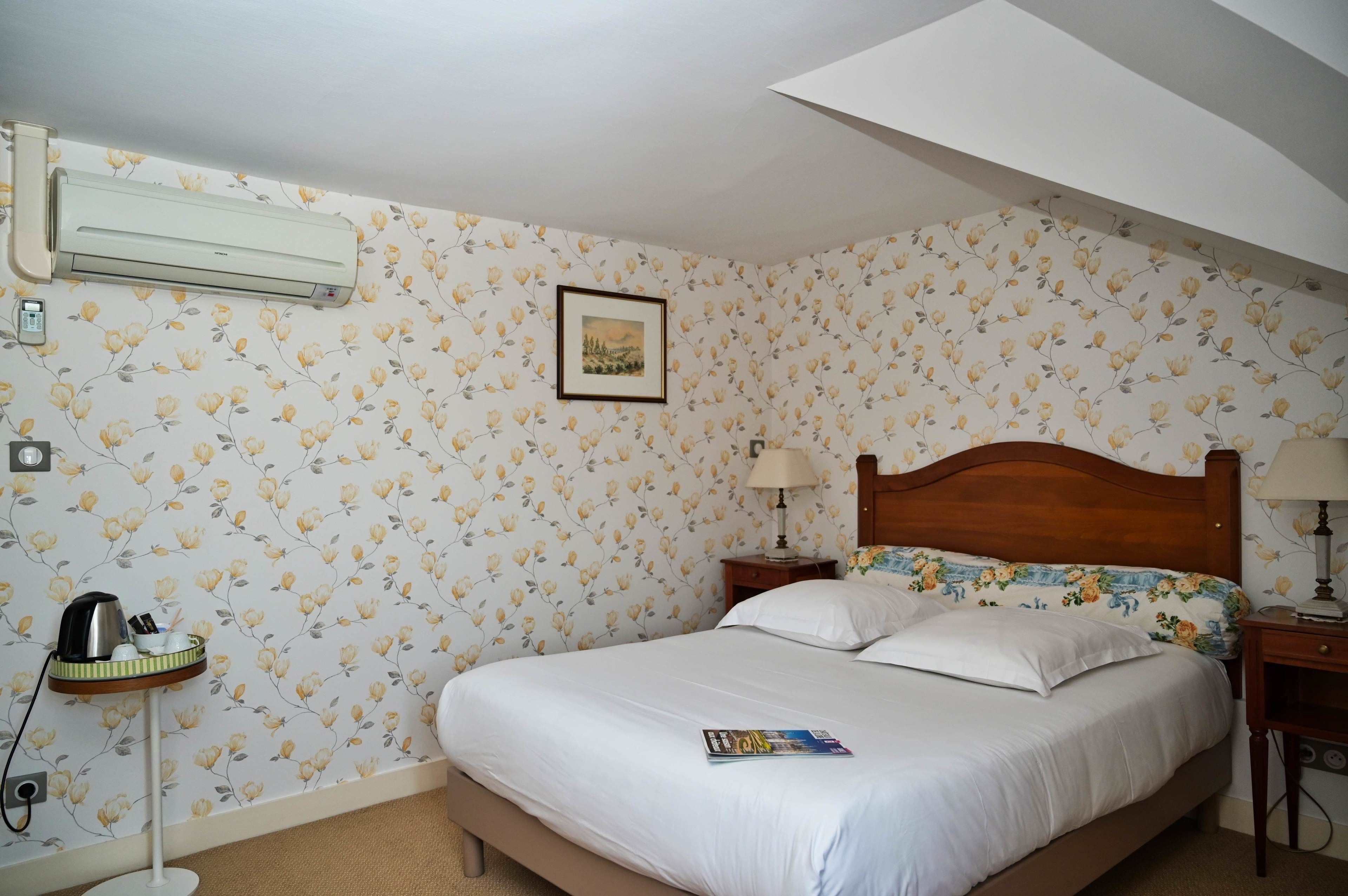A bedroom with a bed, a nightstand, a lamp, an air conditioner, and a floral wallpaper.