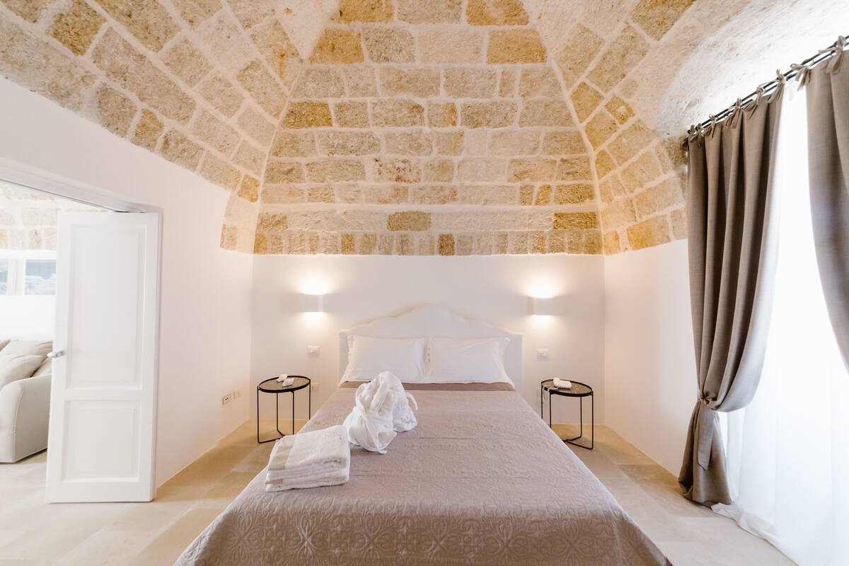 A room with a stone ceiling, a bed with white linens, two bedside tables, and a window with curtains.