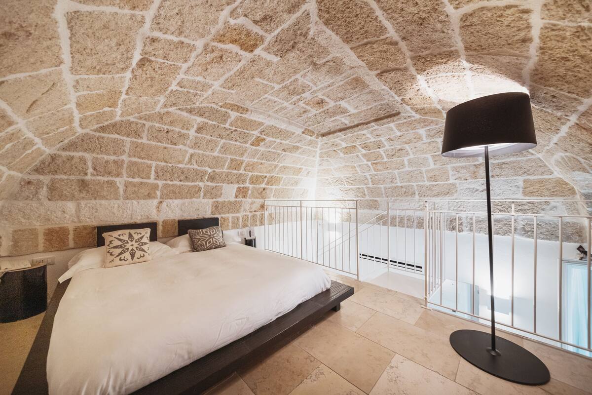 A bedroom with a stone ceiling, a bed with white bedding, a black floor lamp, and a balcony with a railing.