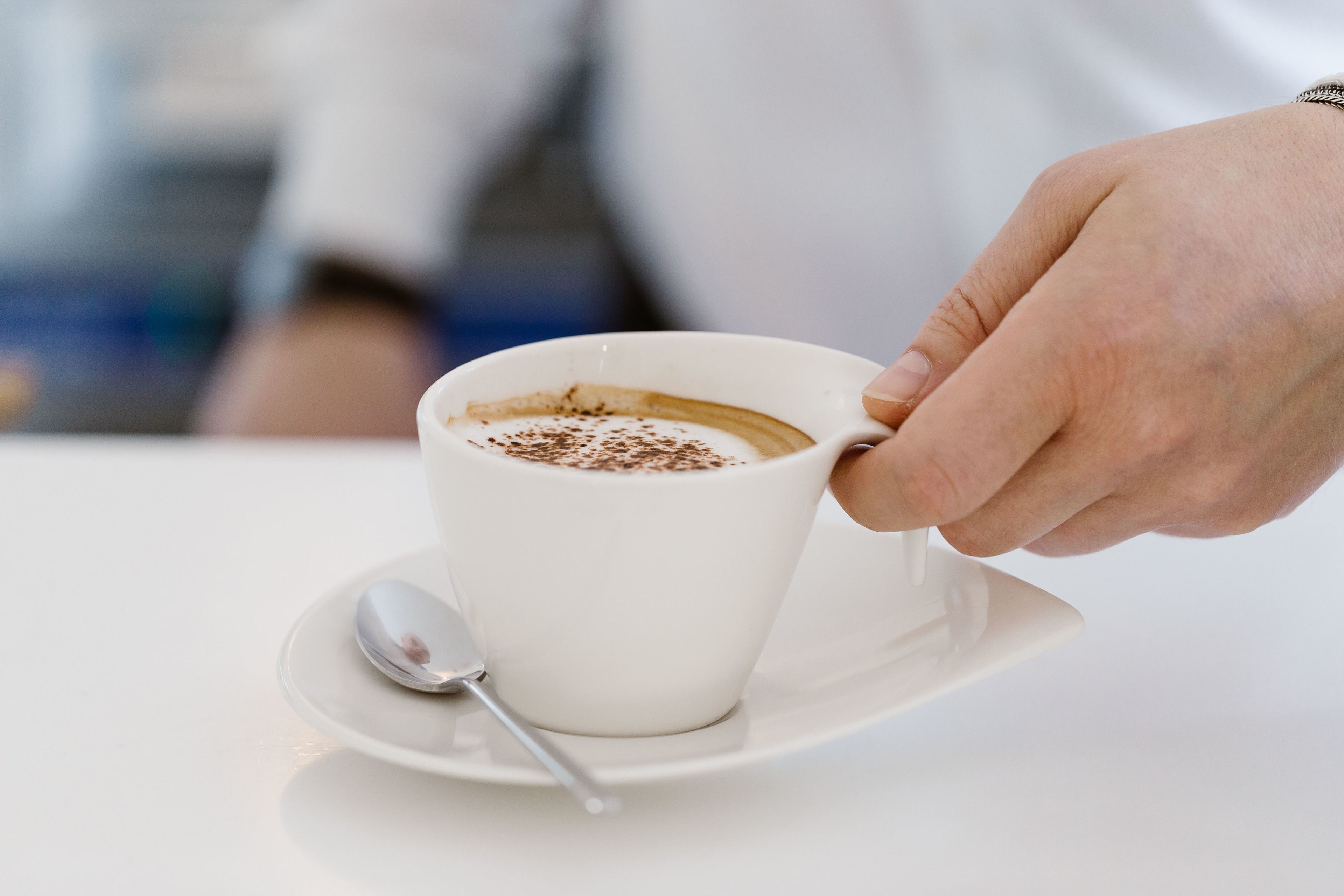 A hand holding a white cup of coffee with a spoon on a saucer.