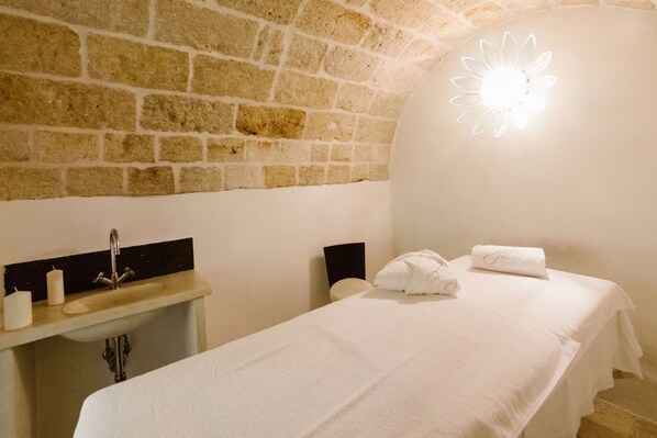 Una habitación con pared de piedra, cama con ropa blanca, lavamanos con grifo y una lámpara de pared.