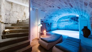 A cozy, stone-walled room with a stone bench, a small table with a lamp, and a view of a stone archway.