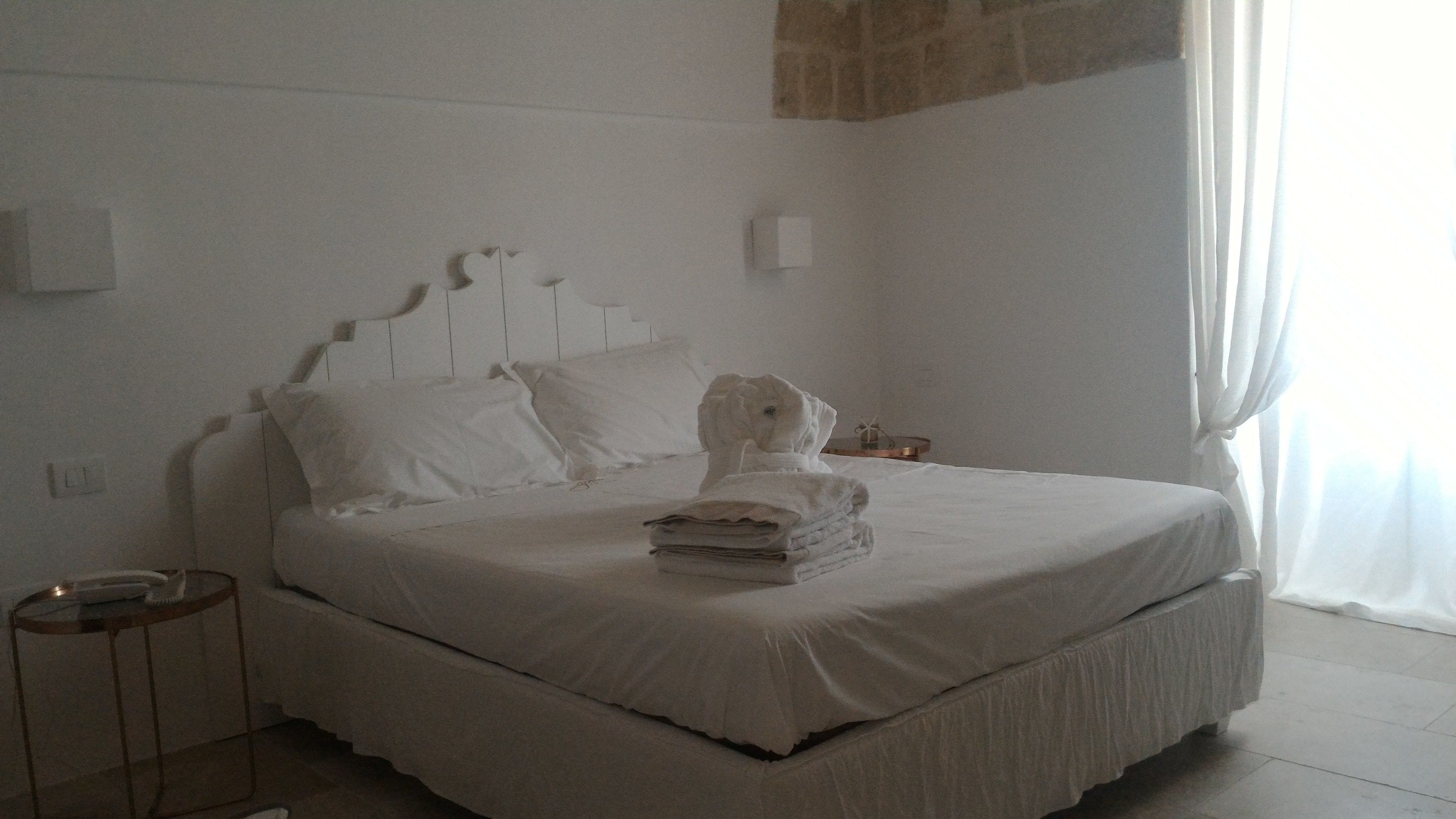 A neatly made bed with white linens and a folded towel on top, a bedside table with a lamp, and a window with curtains.