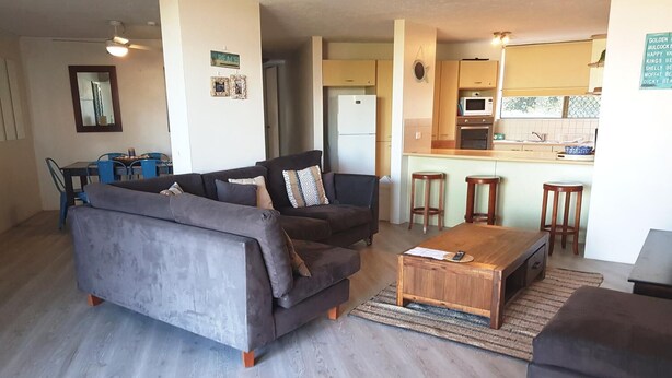 A living room with a grey sofa, a wooden coffee table, and a kitchen area with a microwave and stools. - Casablanca Beachfront Holiday Apartments (Kings Beach)
