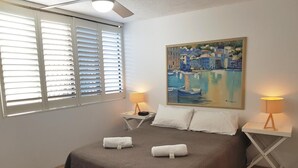 A bedroom with a bed, two bedside tables with lamps, a painting on the wall, and a window with blinds. - Casablanca Beachfront Holiday Apartments (Kings Beach)