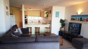 A modern living room with a grey sofa, a TV, and a kitchen in the background. - Casablanca Beachfront Holiday Apartments (Kings Beach)