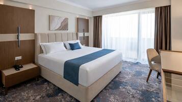 Habitación de hotel con una cama grande, un escritorio y una silla. Se observa un mesita de noche con un teléfono y una lámpara. La habitación presenta un diseño moderno, con un ventanal y cortinas.