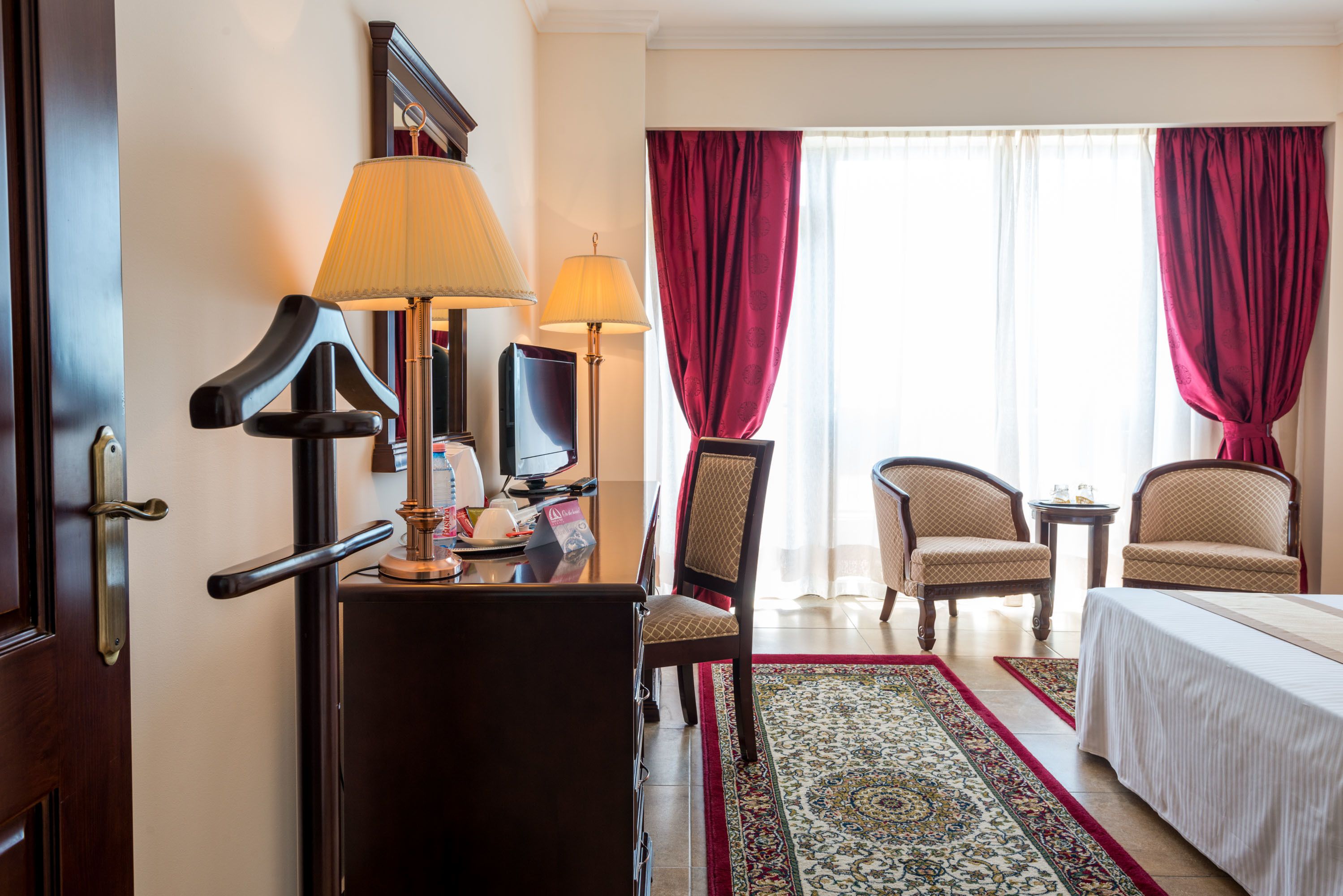 Ein Hotelzimmer mit einem Bett, zwei Sessel, einem Schreibtisch mit Lampe, einem Fernseher und einem großen Fenster mit Vorhängen.