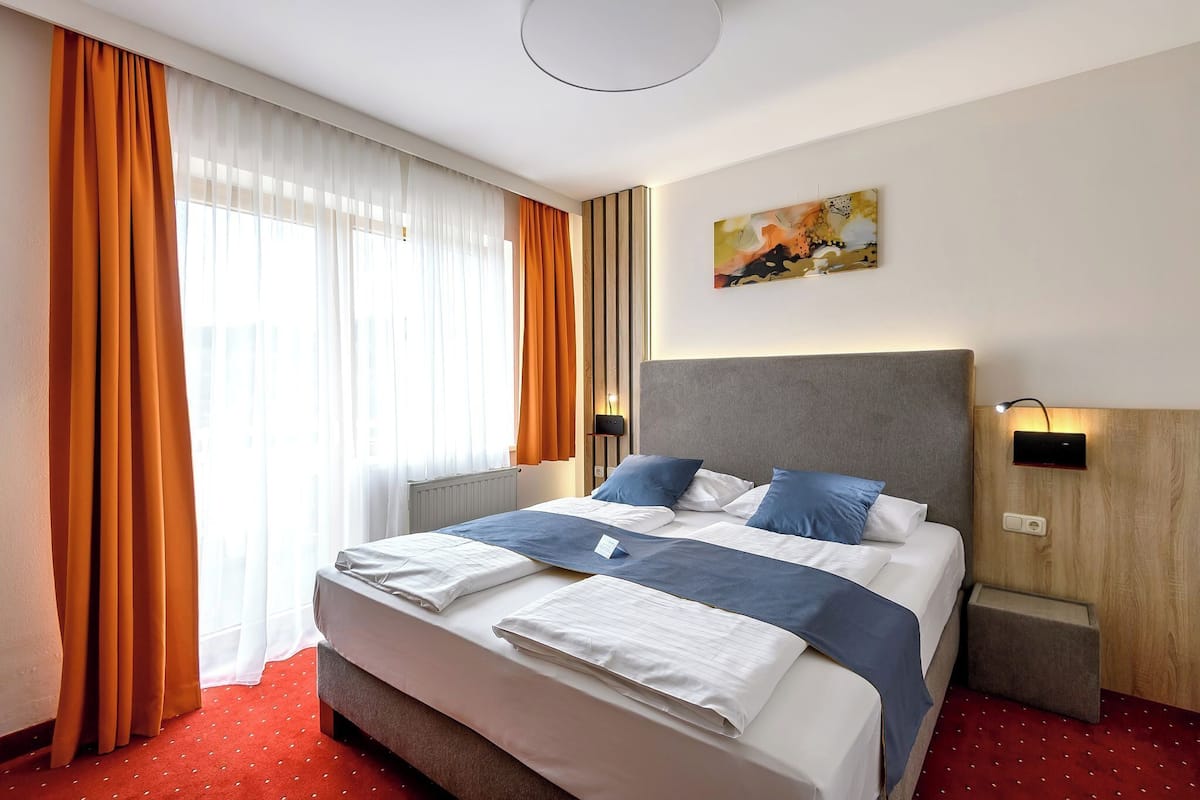 A hotel room with a large bed, two bedside tables, a nightstand, a wall-mounted lamp, and a window with curtains.