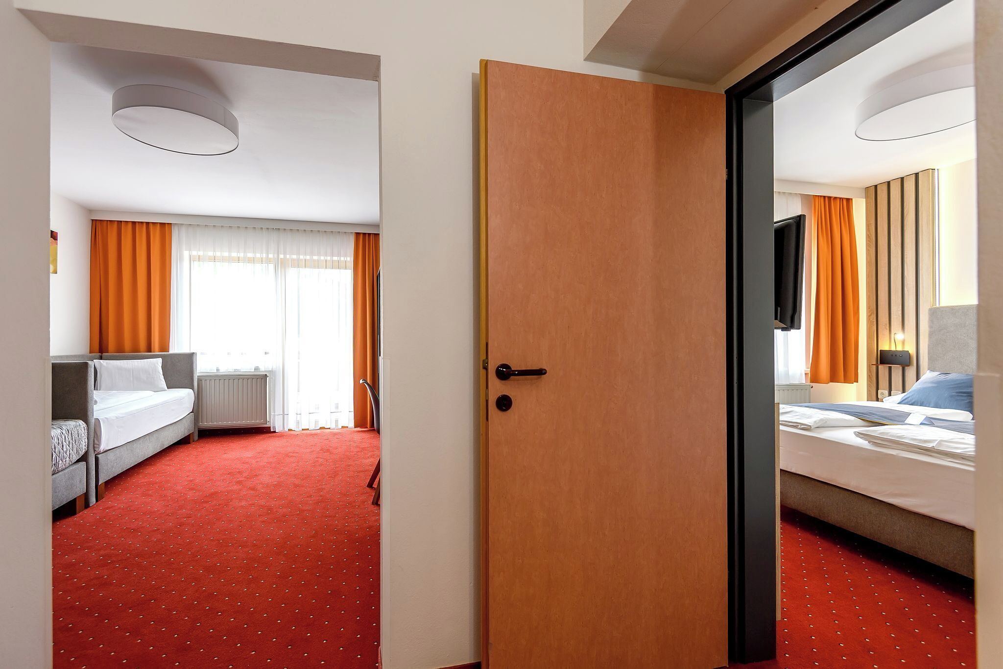 A hotel room with a red carpet, a bed with white linens, an orange curtain, and a television on the wall.