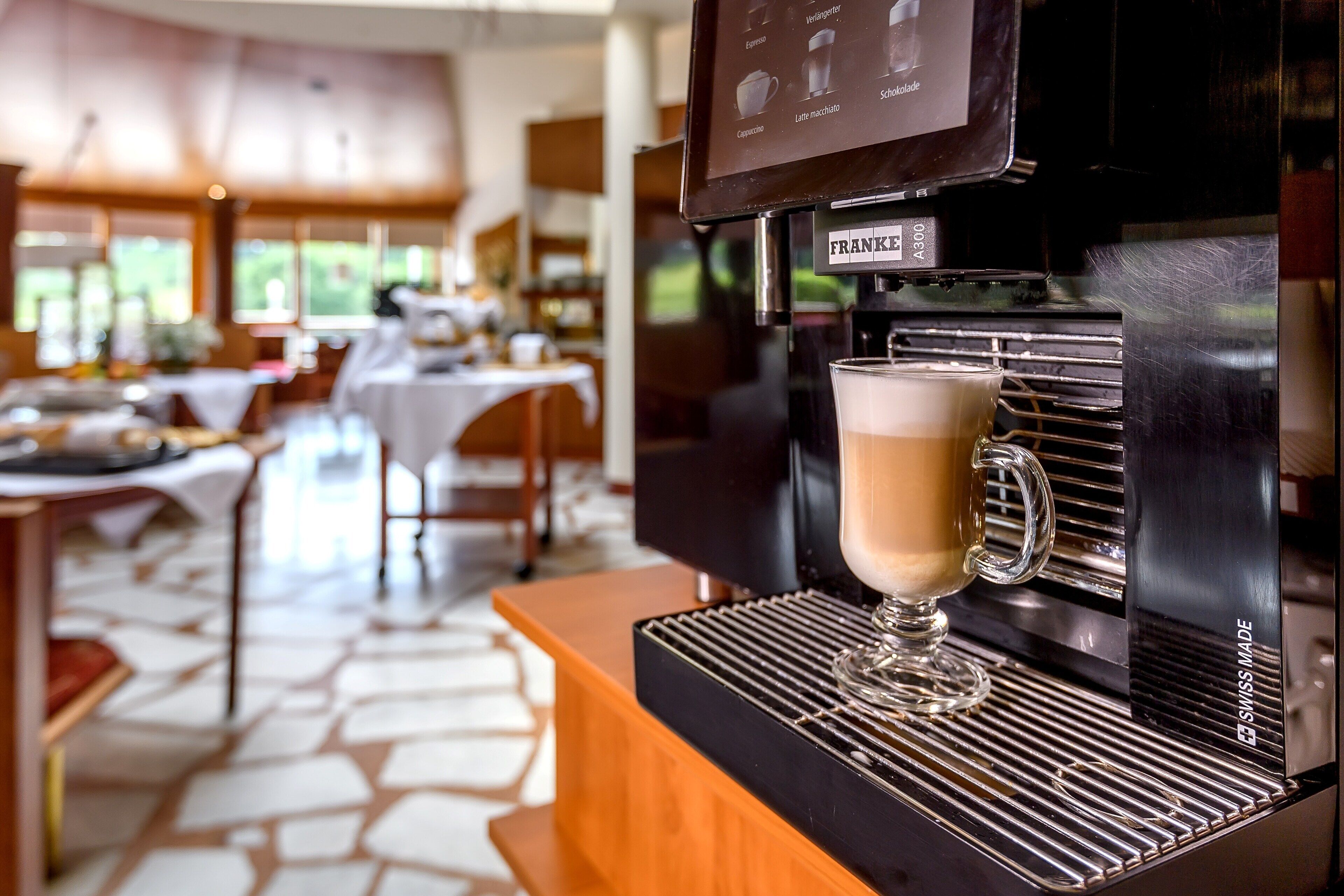 Una cafetera con una taza de café, un menú colgando arriba y un comedor al fondo.