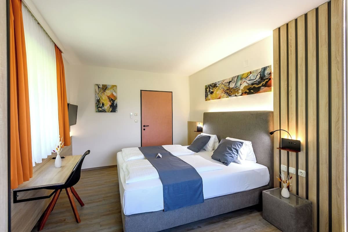 A hotel room with a bed, bedside table, lamp, artwork, and a door.