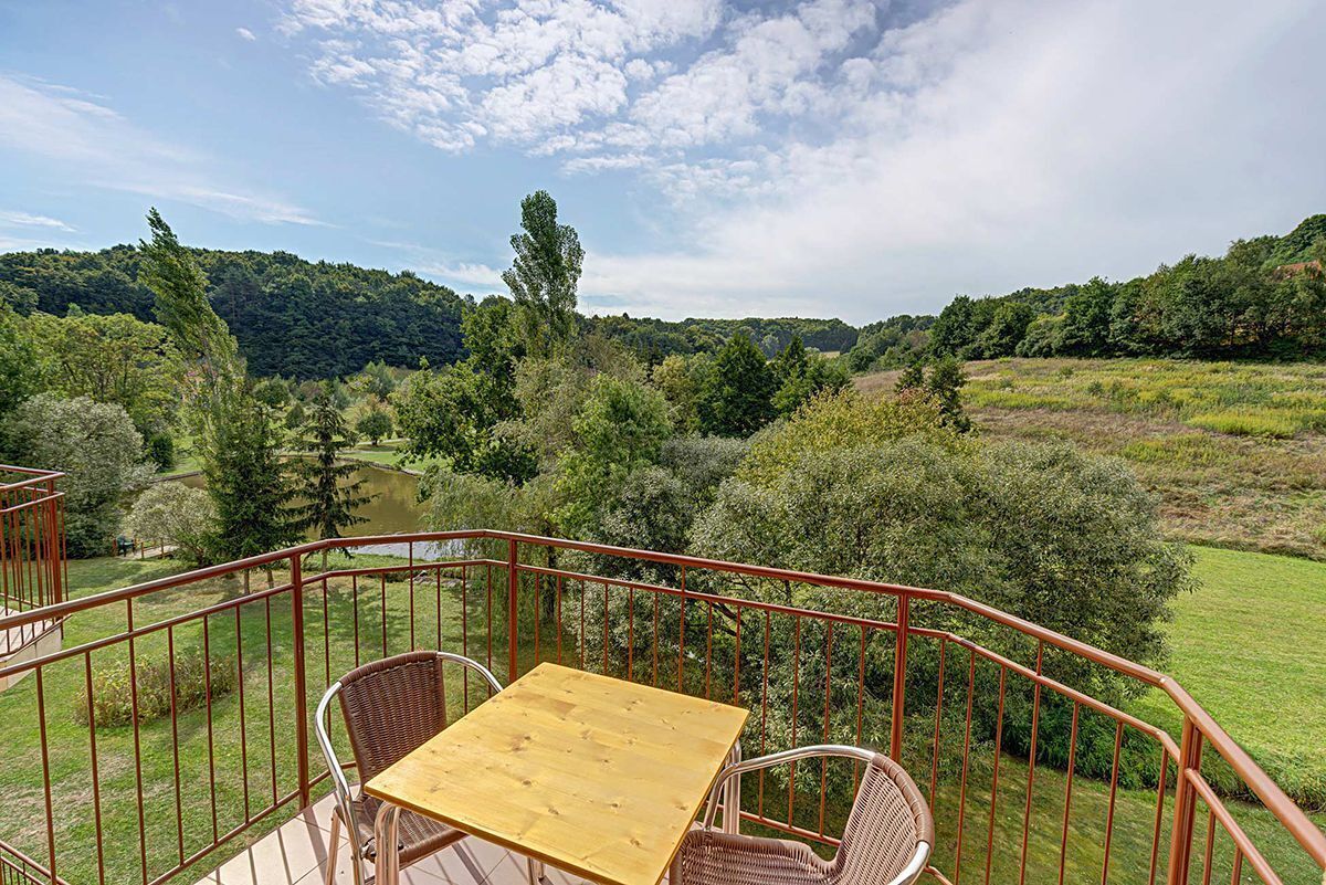 A balcony with a wooden table and wicker chairs, overlooking a lush landscape with trees and a hill in the distance.
