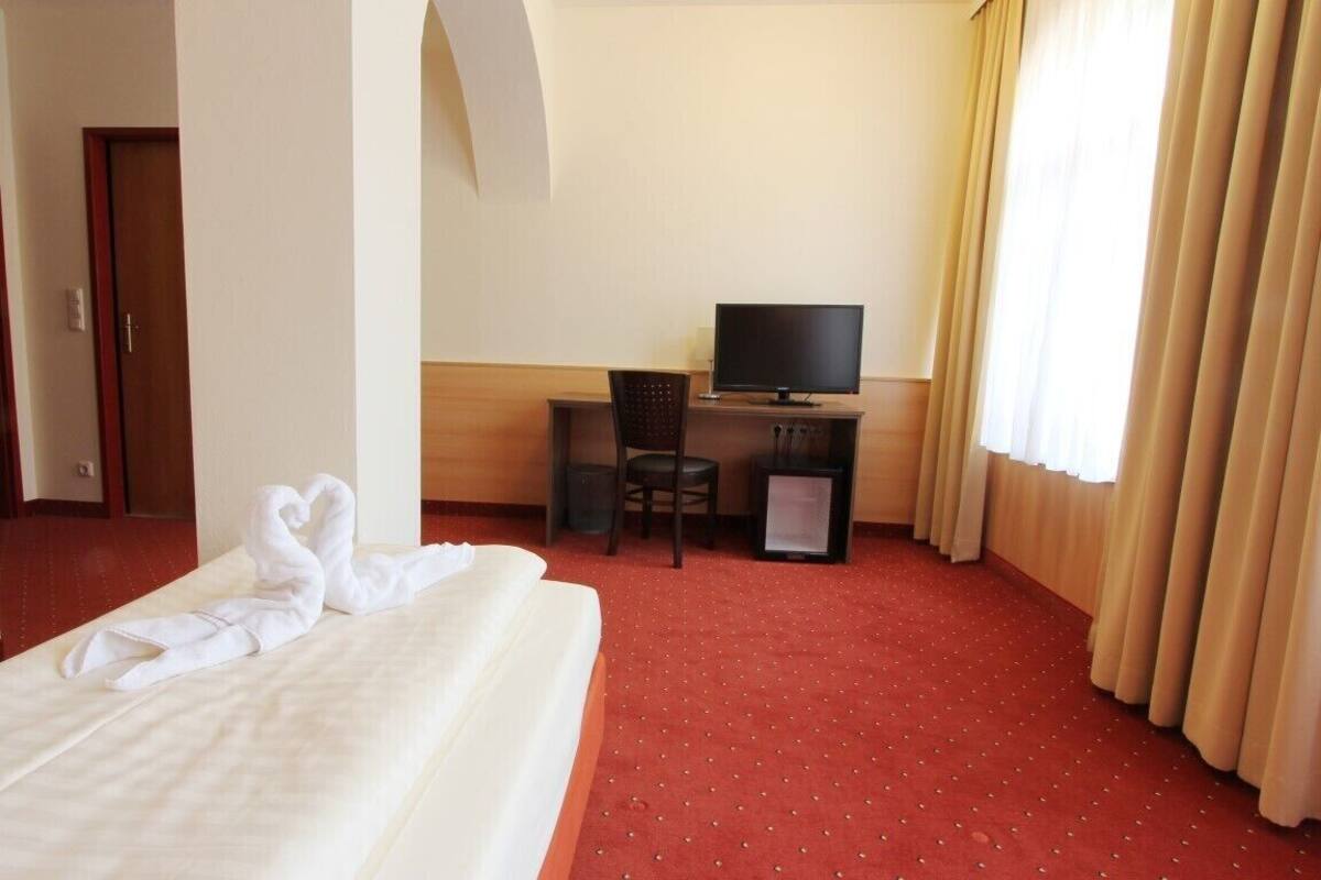 A hotel room with a bed, a desk with a computer and chair, a television, and a window with curtains.