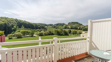 Un balcón con barandilla blanca, una mesa de vidrio y vistas a un paisaje verde con árboles y casas.