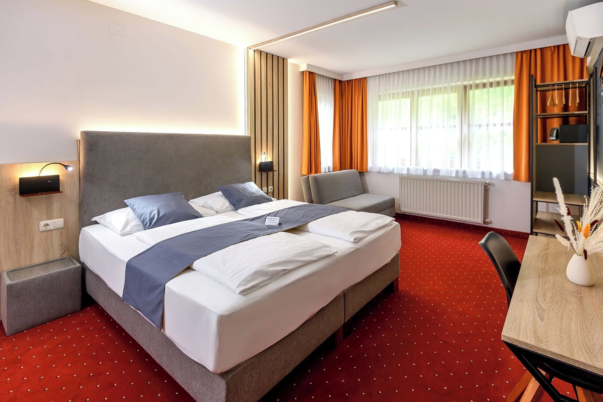 A hotel room with a large bed, a desk with a chair, a radiator, and a window with curtains.