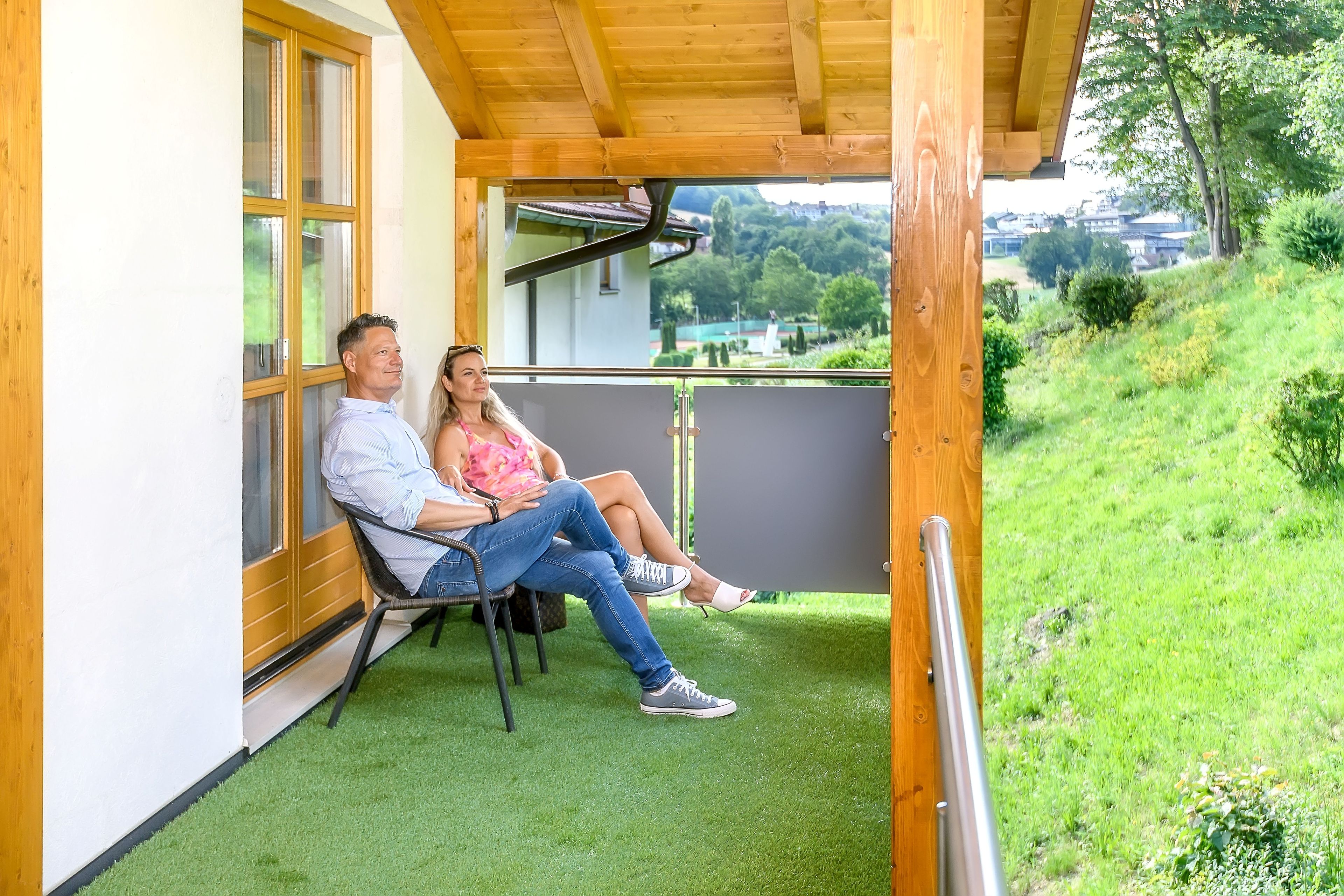Ein Paar sitzt auf einer Bank in einem überdachten Außenbereich mit künstlichem Rasen und blickt auf einen angelegten Garten sowie auf Gebäude in der Ferne.