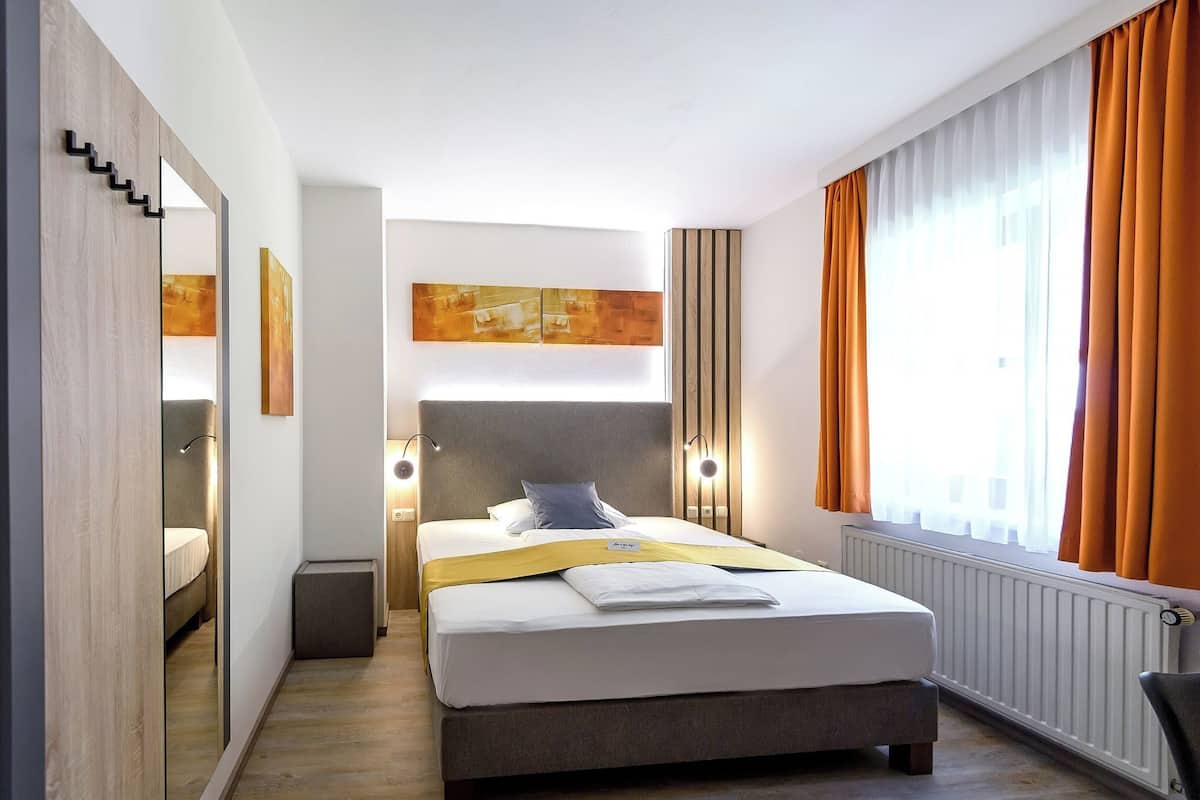 A modern hotel room with a large bed, a mirror, a headboard with integrated lighting, a view of the city through large windows, and a small side table.