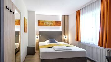 Ett modernt hotellrum med en stor säng, en spegel, en sänggavel med inbyggd belysning, utsikt över staden genom stora fönster och ett litet sidobord.
