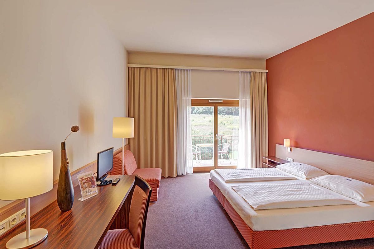 A hotel room with a desk, two beds, a TV, and a balcony with curtains.