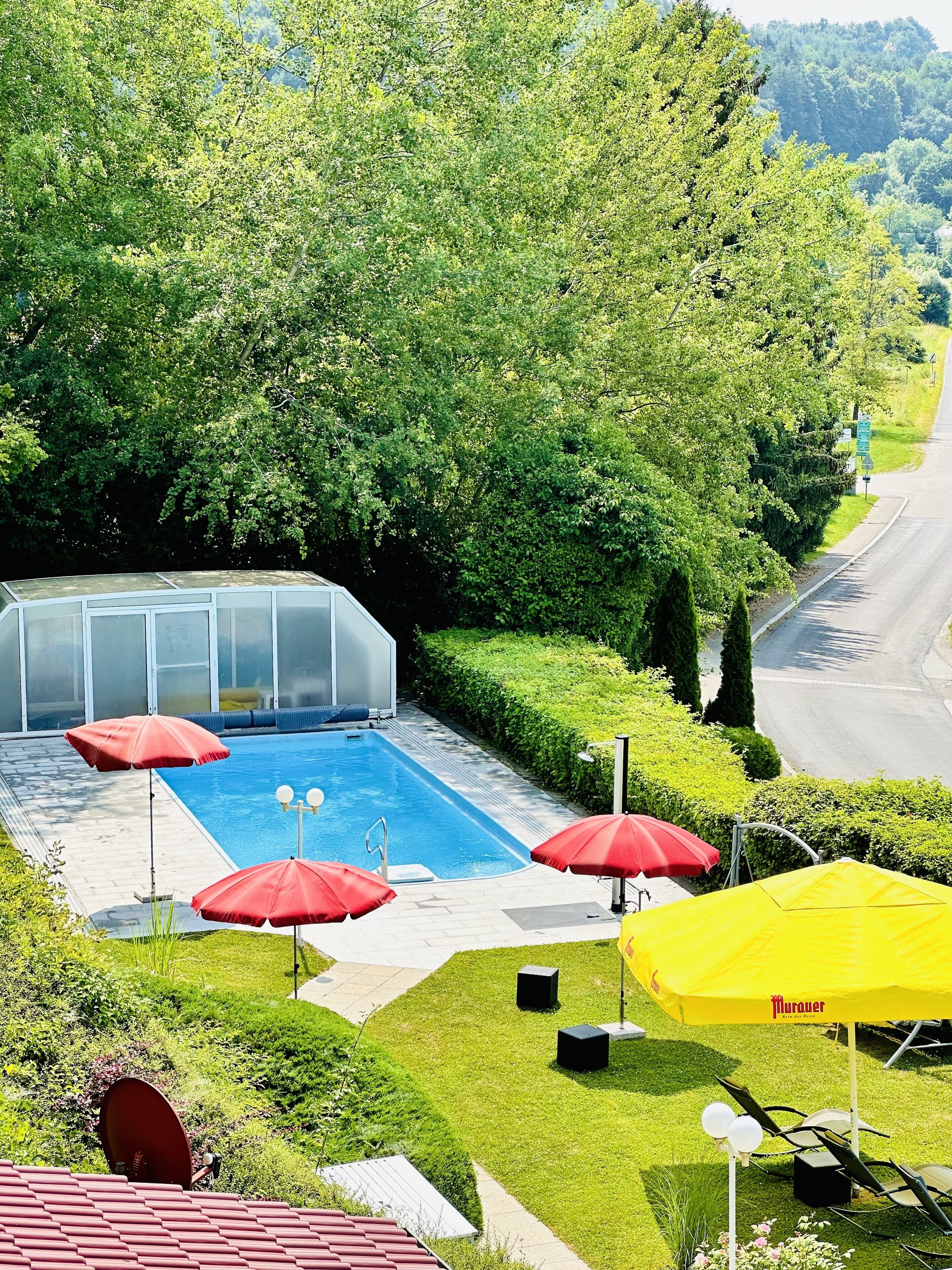 Una piscina con sombrillas rojas y amarillas, rodeada de exuberante vegetación y una carretera al fondo.